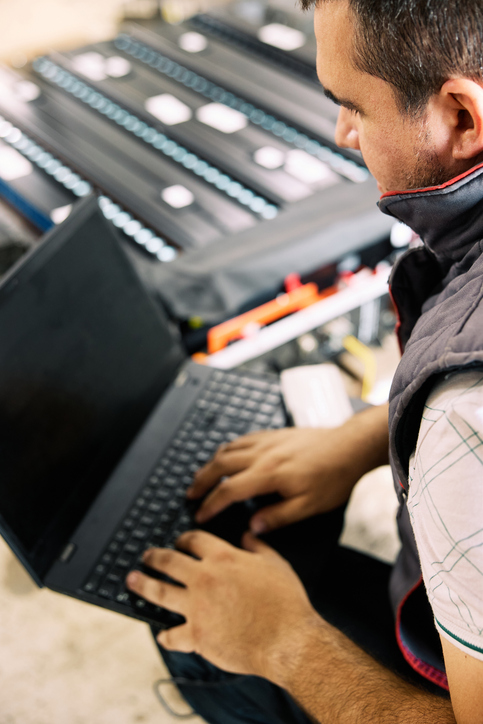 Man working on a laptop beside industrial equipment, inspecting electrical systems and batteries in a workshop